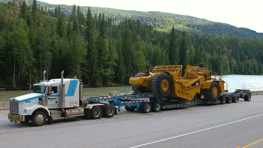 Heavy equipment transport shown by 8 axle RGN with stinger trailer loaded with large CAT oversize overweight scraper. Logi Transports